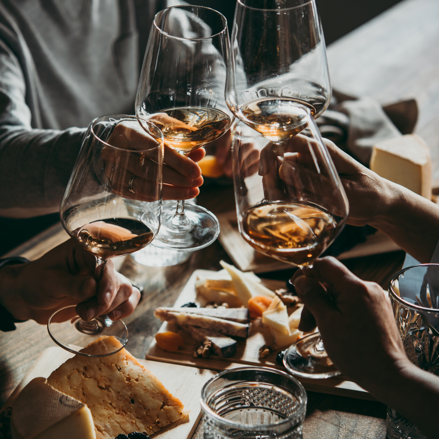 Two people clinking glasses of white wine above a spread of cheese and charcuterie.