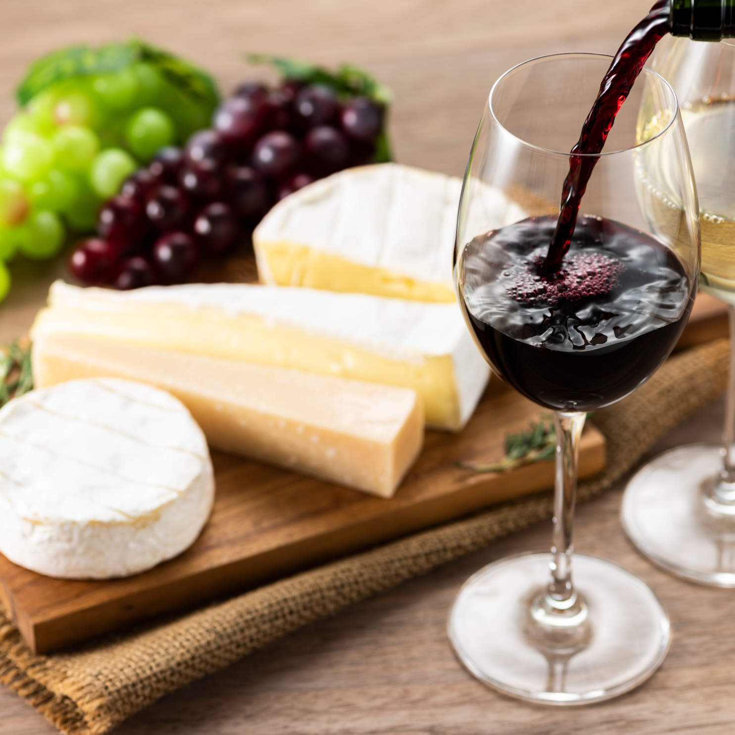 A pour of red wine into a glass beside a wooden board of brie, hard cheese and grapes.